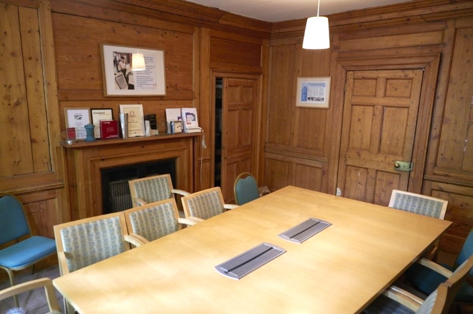 A meeting room in a period property. The walls are panelled in medium-brown wood, the is a fireplace to one side with various papers and awards displayed on top, and a large 8-seater table in the centre.