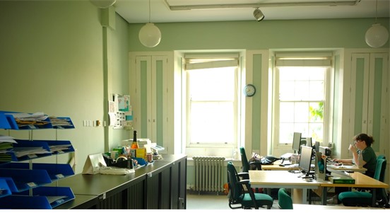 An office space in a period property with pale green walls, and matching green and white panelling. Light shines through two large windows, and a person sips their tea while looking at a computer screen at one of the desks.