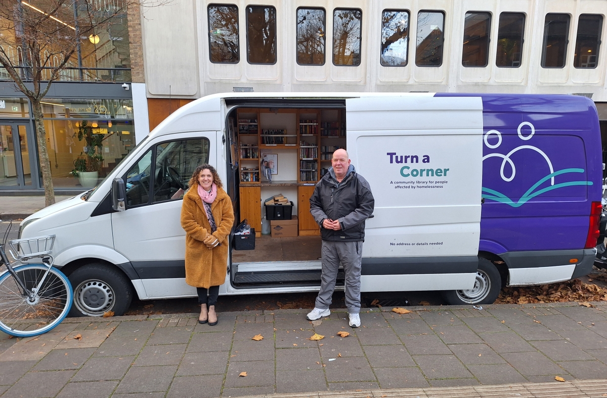 A man and woman stand smiling in front of a white, purple and green mobile library van parked on a city street. On the van is written 'Turn a Corner', and the side door is open to show bookshelves inside.
