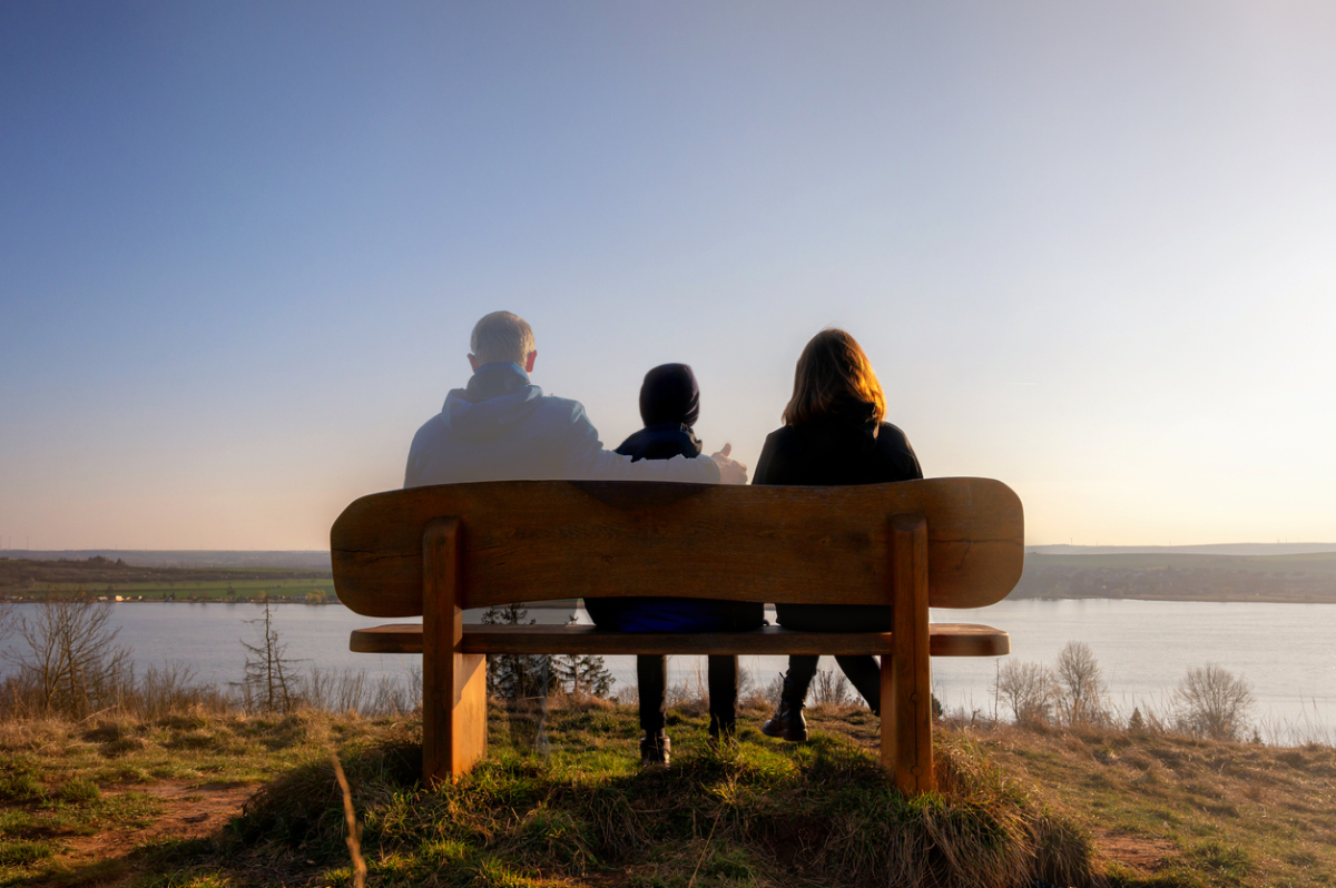 A man, child and woman sit on a wooden bench, photographed from behind. They are on the bank of a lake in front of them, which stretches the width of the photo. The man has his arm around the child, but the man is partially greyed out, as if he is not entirely there.