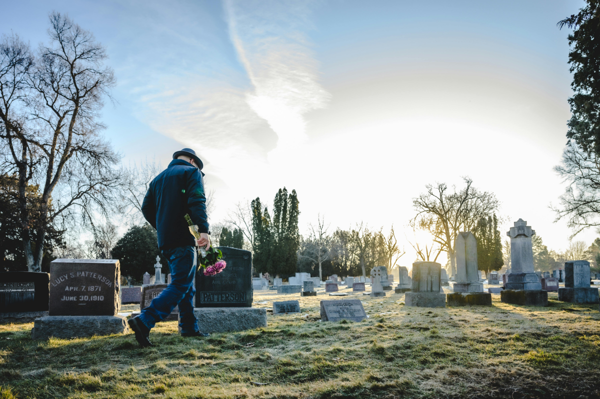 A man is shown from the side, holding pink flowers and walking in a graveyard.
